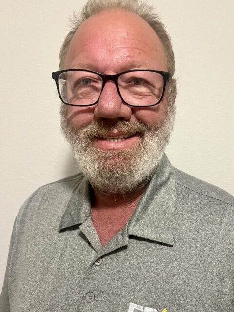A smiling man with glasses and a gray beard wears a gray polo shirt with an EPIC Service Equipment Solutions logo, standing in front of a plain, light-colored wall.