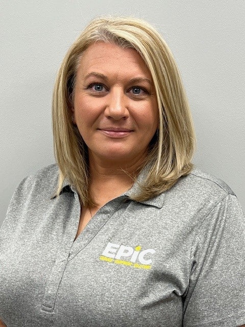 A woman with straight blonde hair in a gray EPIC logo collared shirt stands before a plain light gray wall, smiling slightly at the camera.