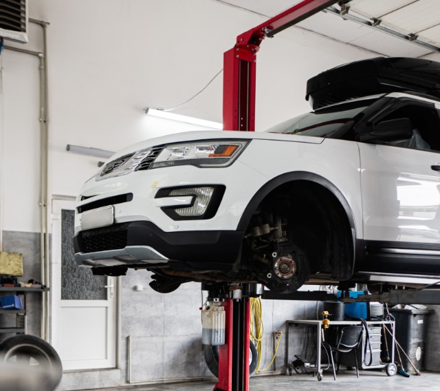 A white SUV with its front wheels removed is elevated on a red hydraulic lift inside an auto repair shop. Tools and equipment are visible in the background.