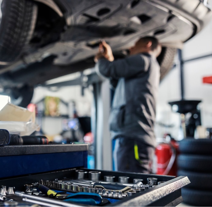 A mechanic works beneath a car on a hydraulic lift, with tools and sockets in the foreground. Mechanic and background are blurred.