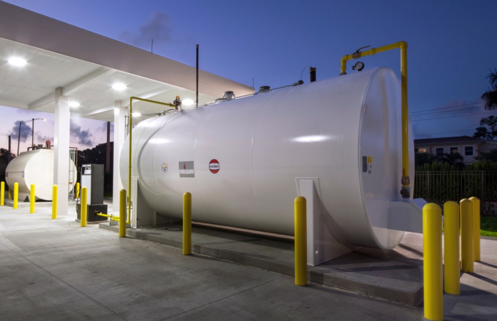 Large white fuel storage tank at a fueling station, yellow safety bollards around it, canopy overhead, another tank in back at dusk.