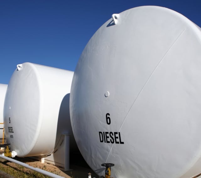 Three large white cylindrical fuel storage tanks labeled DIESEL sit outdoors under a clear blue sky. The tank in the foreground is marked with the number 6.