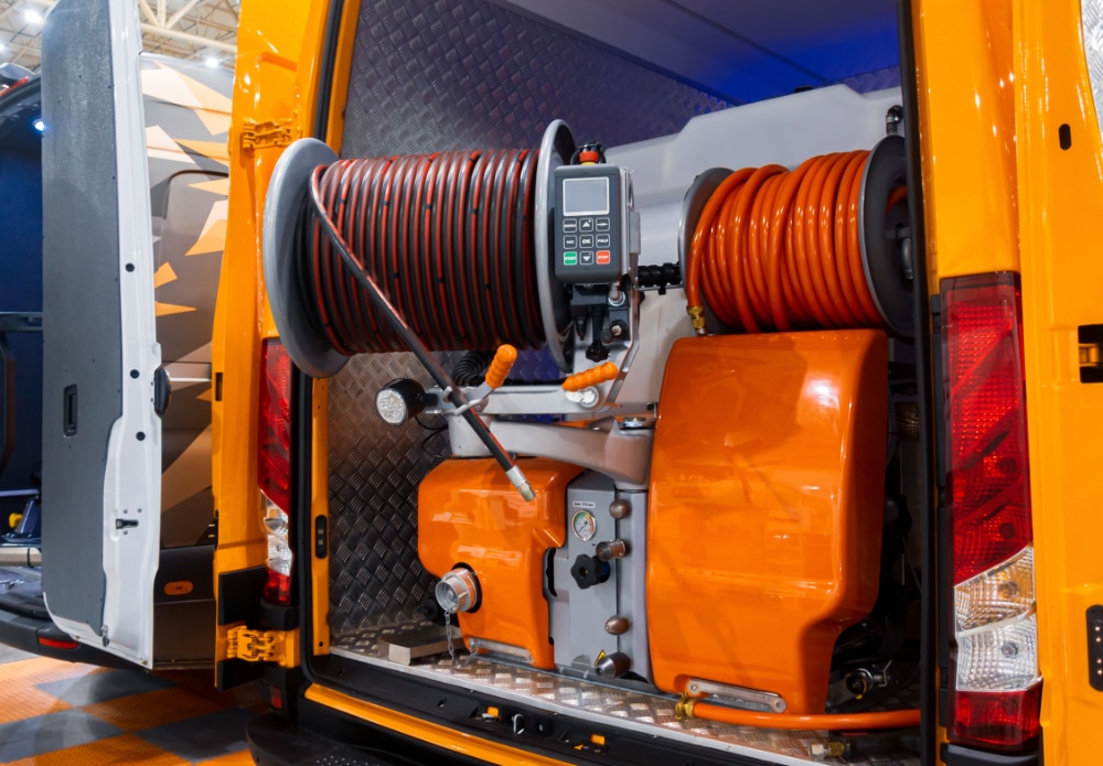 Back of yellow utility van open, showing orange hoses on reels, a control panel, and assorted valves and levers for maintenance.