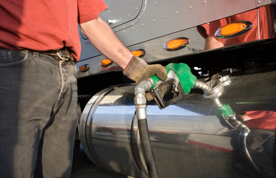 A gloved person refuels a large truck with a green nozzle in its shiny metallic tank; the truck's side and lights are visible.
