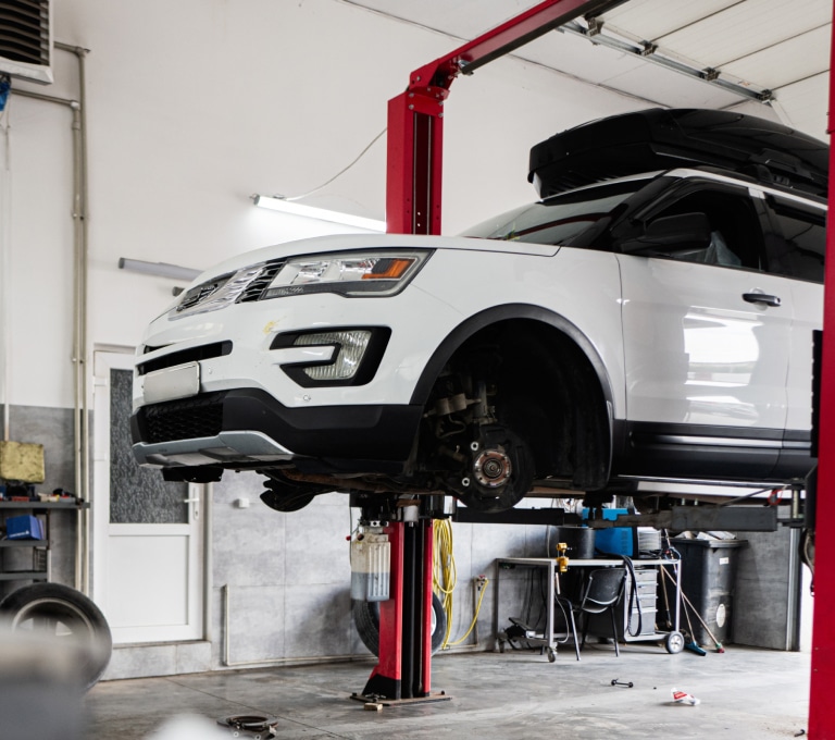 A white SUV with its front wheels removed is lifted on a red hydraulic lift in an auto shop, with tools and equipment in the background.