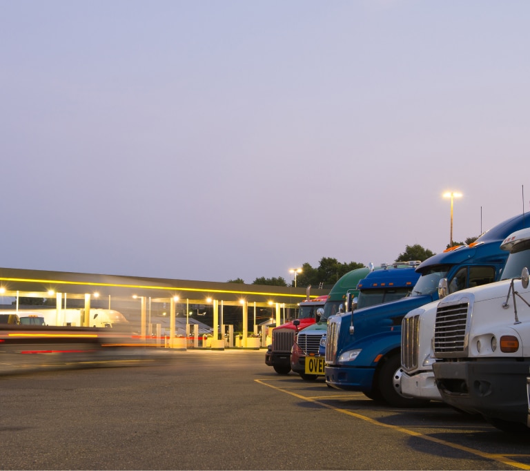 Semi-trucks parked in a row at a brightly lit truck stop in the evening, with clear sky and trees visible in the background.