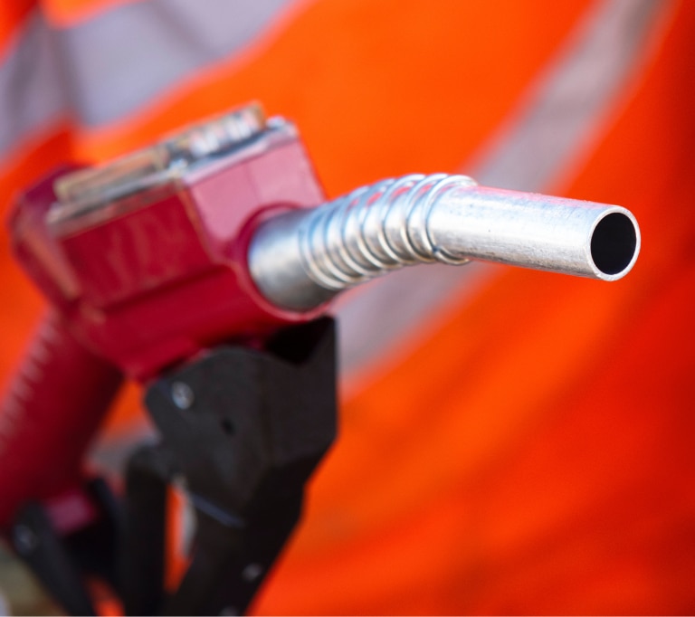 A close-up of a fuel pump nozzle with a red handle, held before a blurred background showing an orange safety vest with reflective stripes.