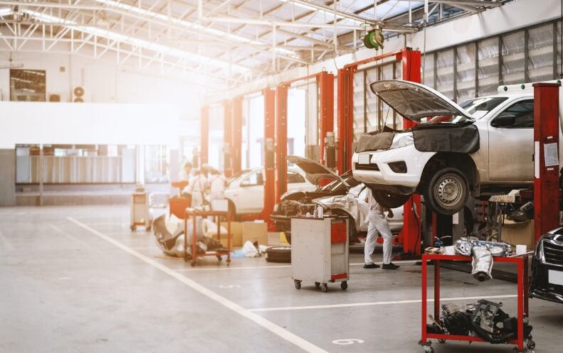 Mechanics working on cars lifted on hydraulic ramps in a spacious, well-lit auto repair shop with tools and car parts organized around the workspace.