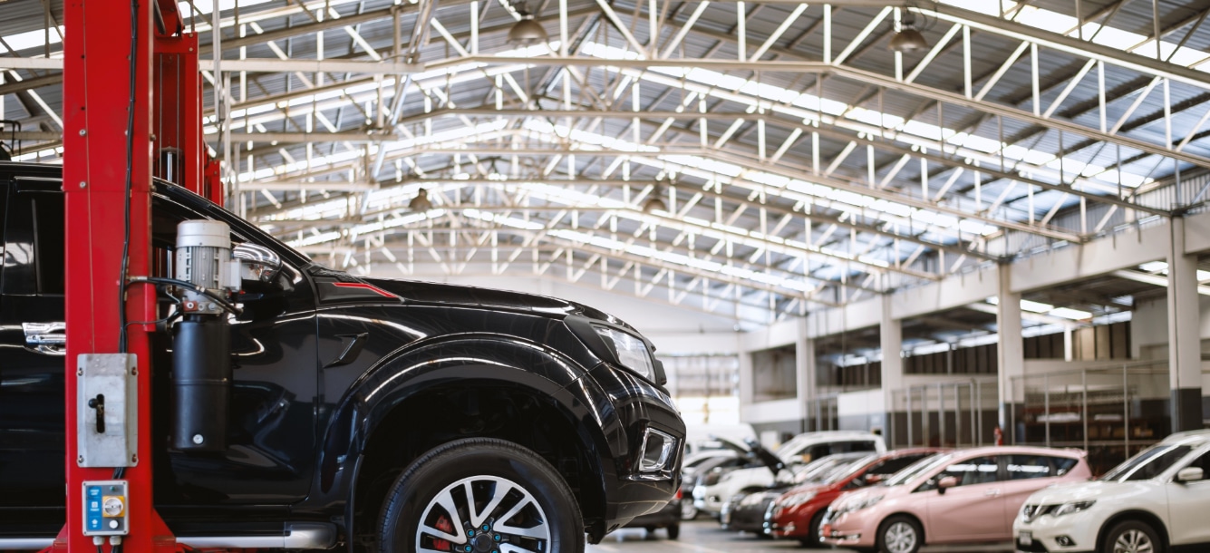 A black vehicle on a hydraulic lift in a spacious, well-lit auto repair shop with multiple cars parked in the background.
