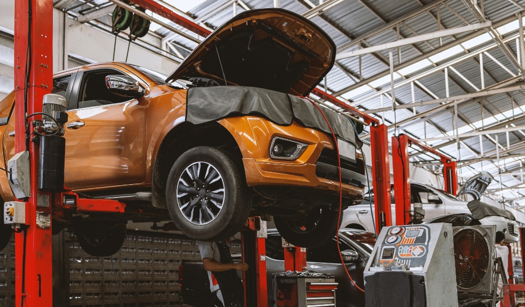 A mechanic works under an orange pickup on a red lift in a bright repair shop, with other vehicles and tools visible behind.
