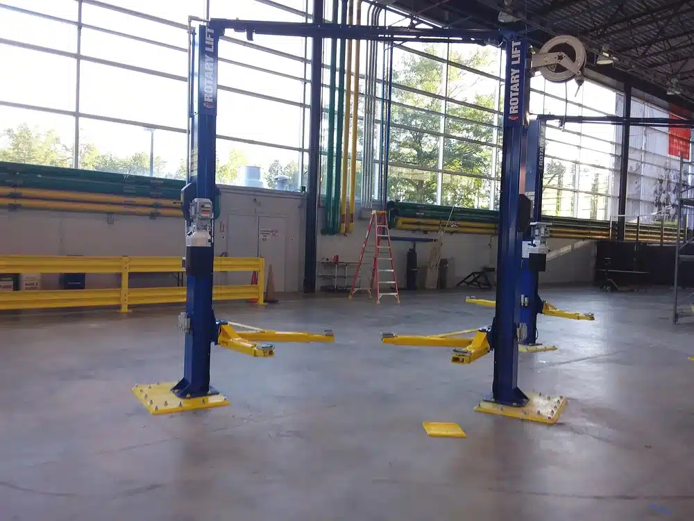 A blue and yellow two-post car lift in a spacious, well-lit garage with large windows, yellow rails, and a red ladder; area is clean.