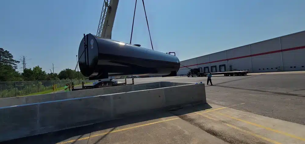 A crane lifts a large cylindrical tank into a concrete enclosure outside an industrial building as workers guide the process nearby.