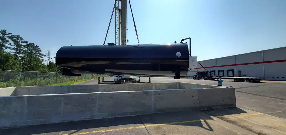 A large black industrial tank is lifted by a crane over a concrete enclosure outside, near a warehouse and trees on a sunny day.