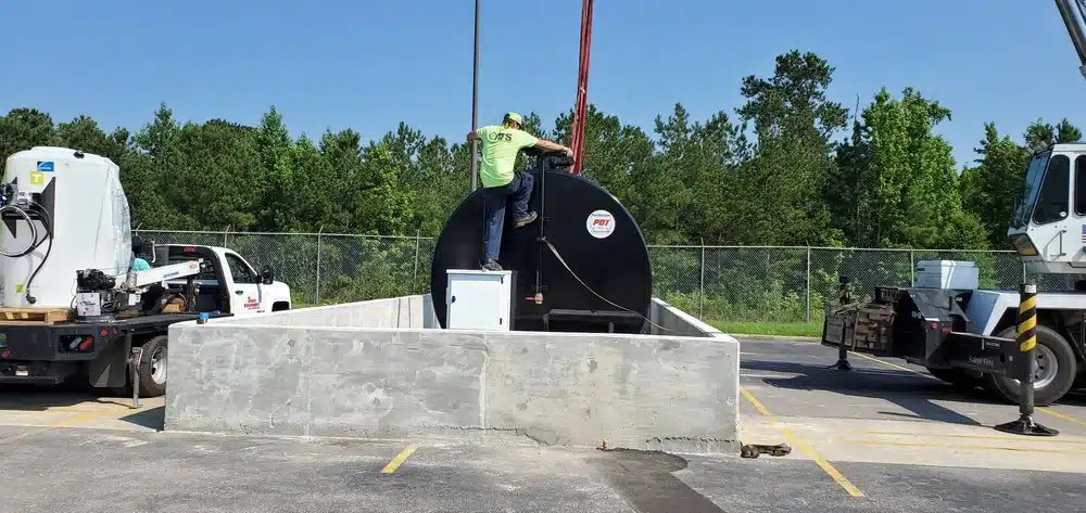 A worker in a green shirt stands on a concrete platform by a large black fuel tank outdoors, with trucks and trees behind him.