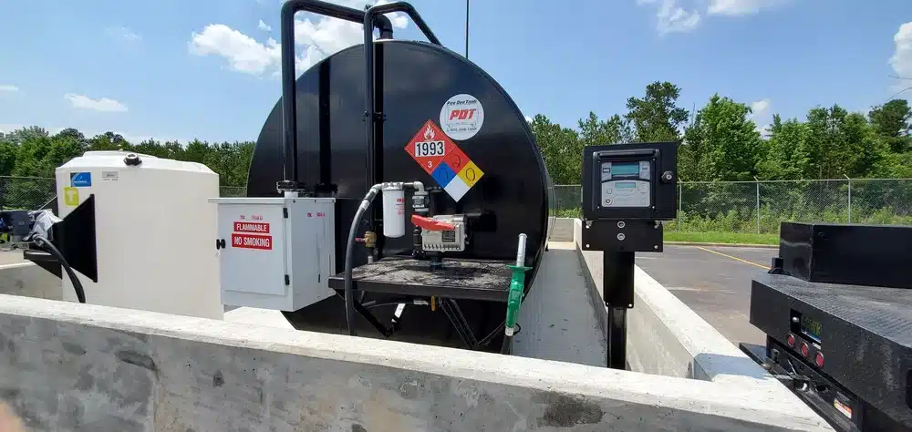 A large black fuel storage tank with hazard labels sits in a concrete enclosure outdoors, near utility boxes and green trees.