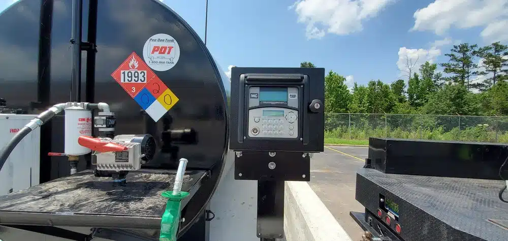 A fuel pump with a digital meter and hazard labels stands outside by a black tank, near trees and a fence under a partly cloudy sky.