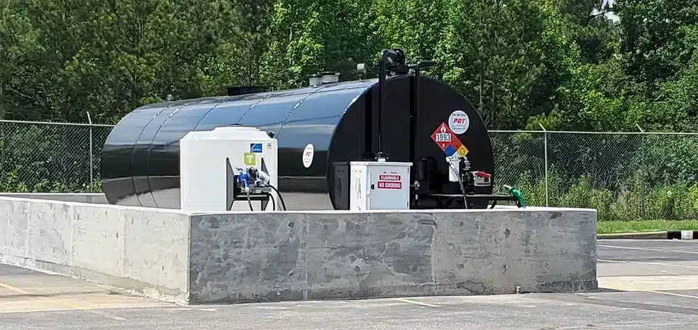 A large black above-ground fuel tank in a concrete containment area, surrounded by trees and fence, with pumps and warning signs.