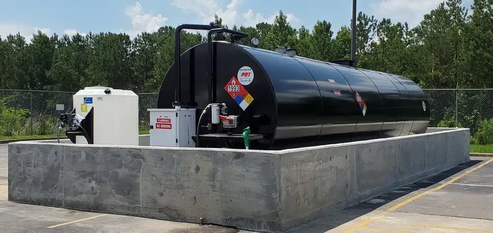 A large black fuel storage tank with hazard labels in a concrete barrier outdoors, near trees, a fence, a white utility box, and tank.