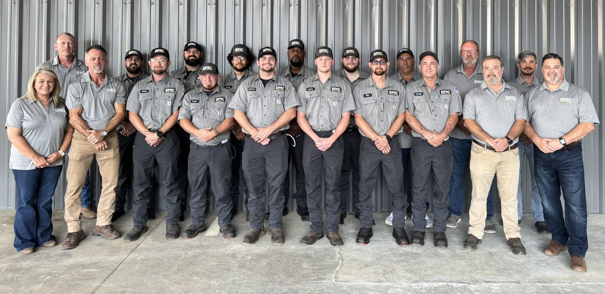Seventeen people in gray shirts and black or blue pants stand indoors before a corrugated metal wall, posing for a group photo.