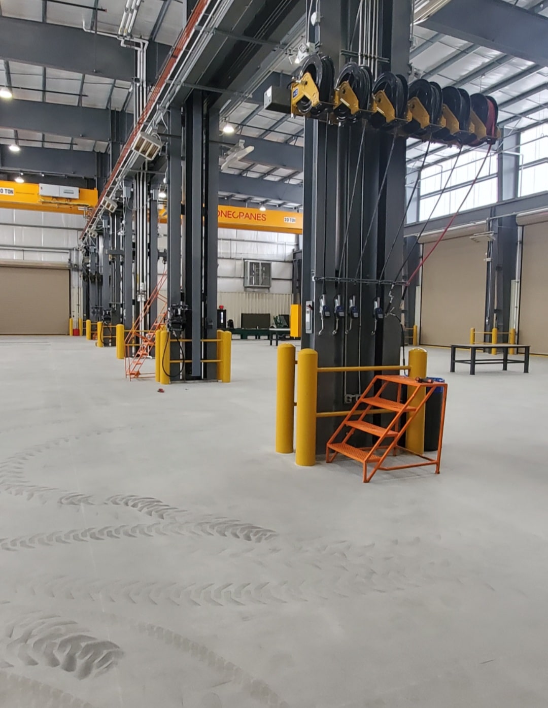 Spacious industrial warehouse with tall black columns, overhead cranes, yellow bollards, retractable hoses, and an orange ladder.