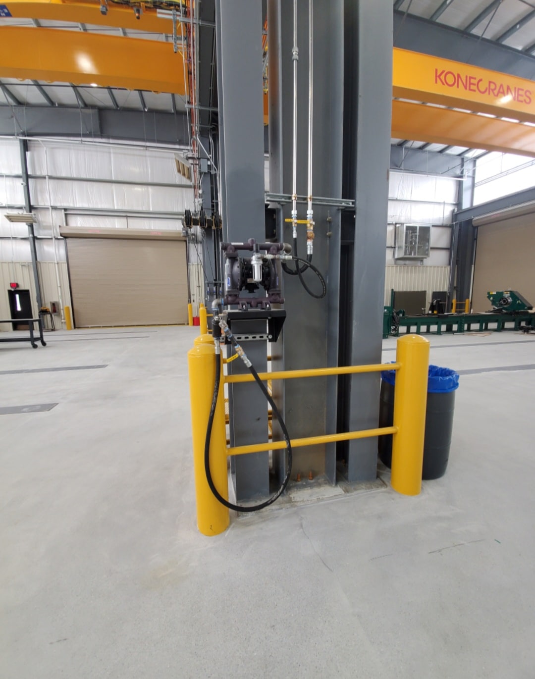 A metal column in an industrial building with pipes, hoses, controls, yellow safety rails, bollards, blue trash bin, and Konecranes crane above.