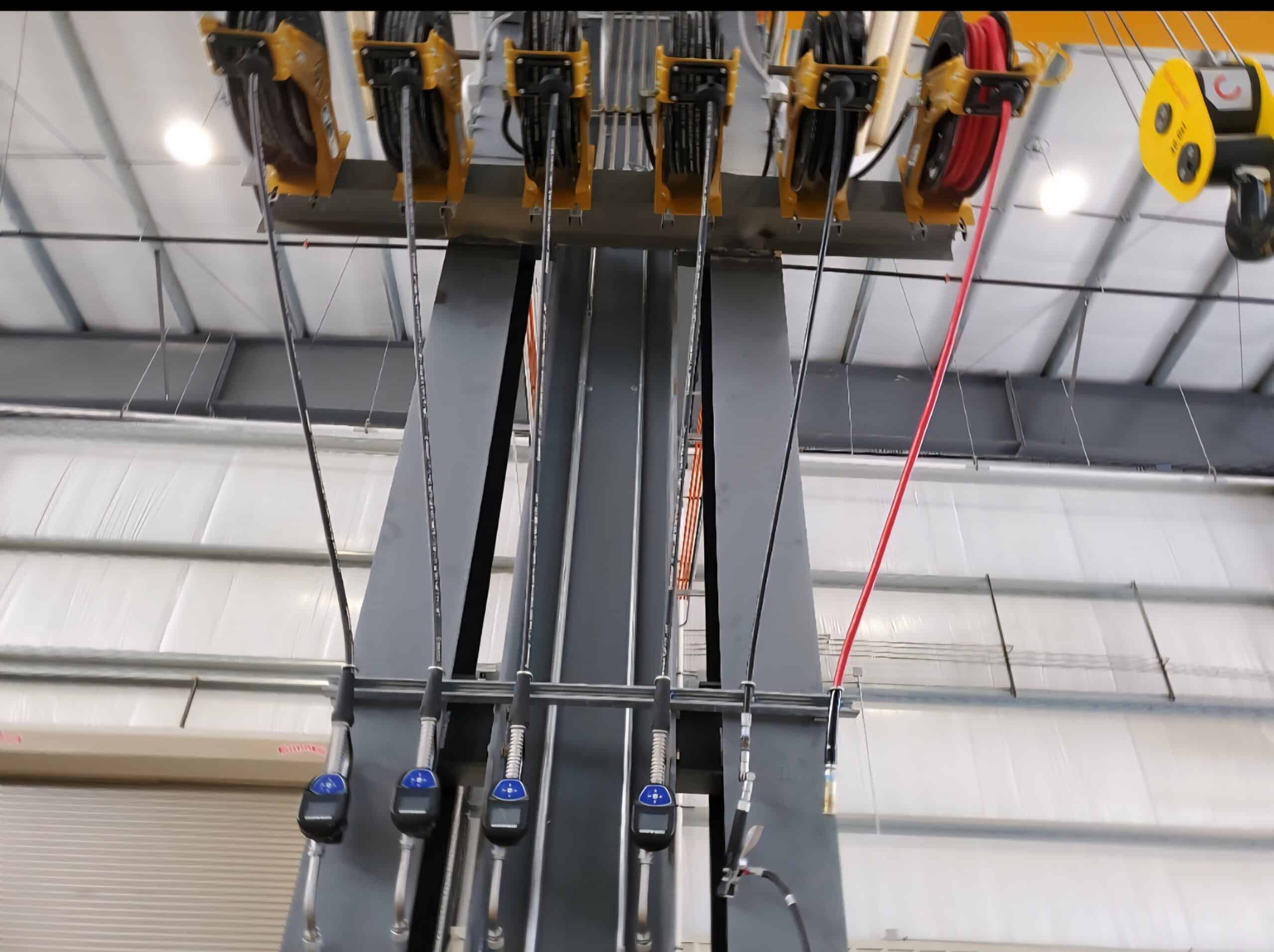 Close-up view of an industrial overhead crane system with steel cables, pulleys, and controls inside a warehouse with a high metal ceiling.