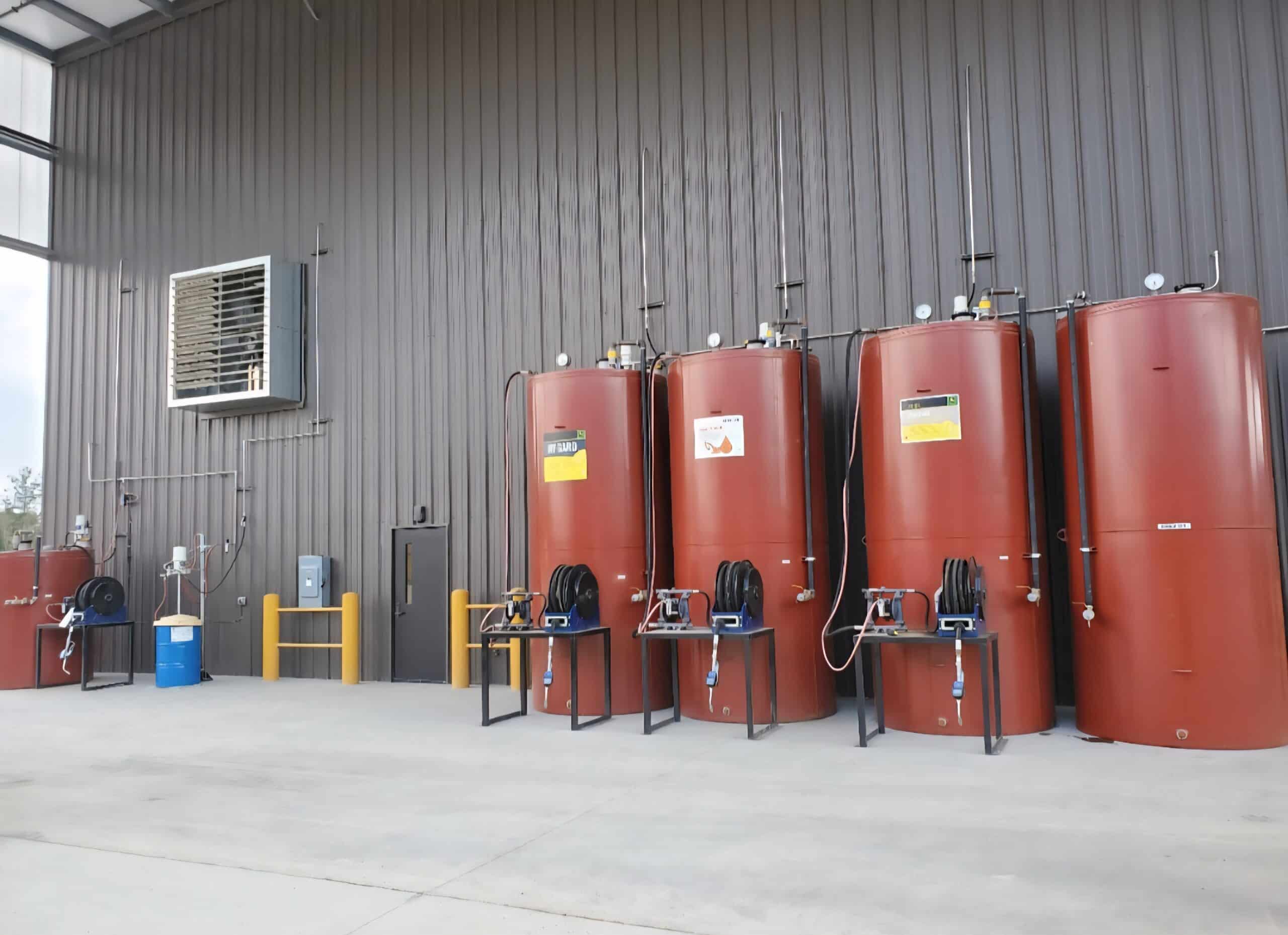 Four large red storage tanks with hoses and meters stand by a gray wall in a warehouse; a blue drum and yellow barriers are on the left.