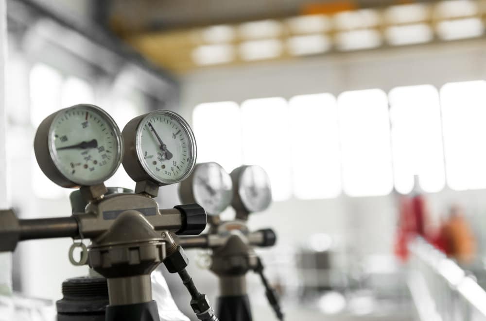 Close-up view of two pressure gauges connected to metal pipes in an industrial area, large windows softly lit in the blurred background.