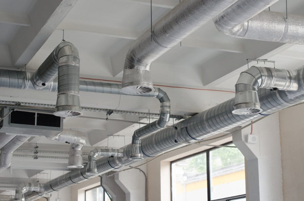 Exposed metal air ducts and round vents hang from the ceiling in an industrial-style space with large windows.