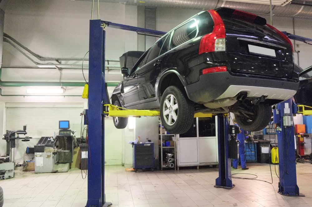 A black SUV on a hydraulic car lift in an auto repair garage with tools, equipment, workstations, and bright lights in the background.
