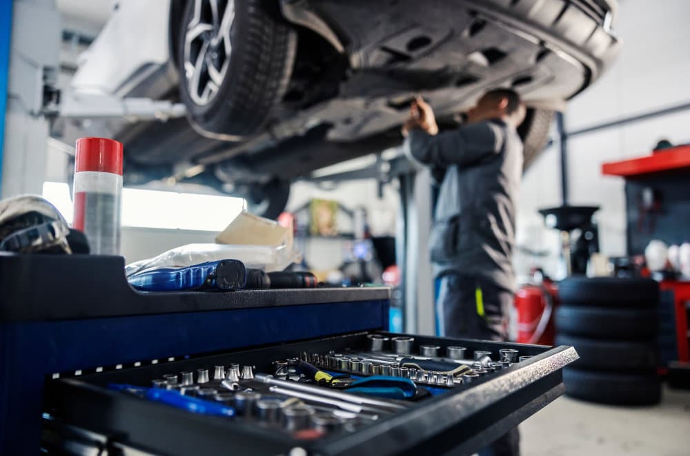 A mechanic works under a lifted car in a garage with an open tool drawer in front showing wrenches, ratchets, and gloves.