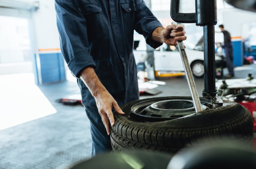 A mechanic in a blue jumpsuit works on a car tire with a tool in an auto repair shop, with a white car in the background.
