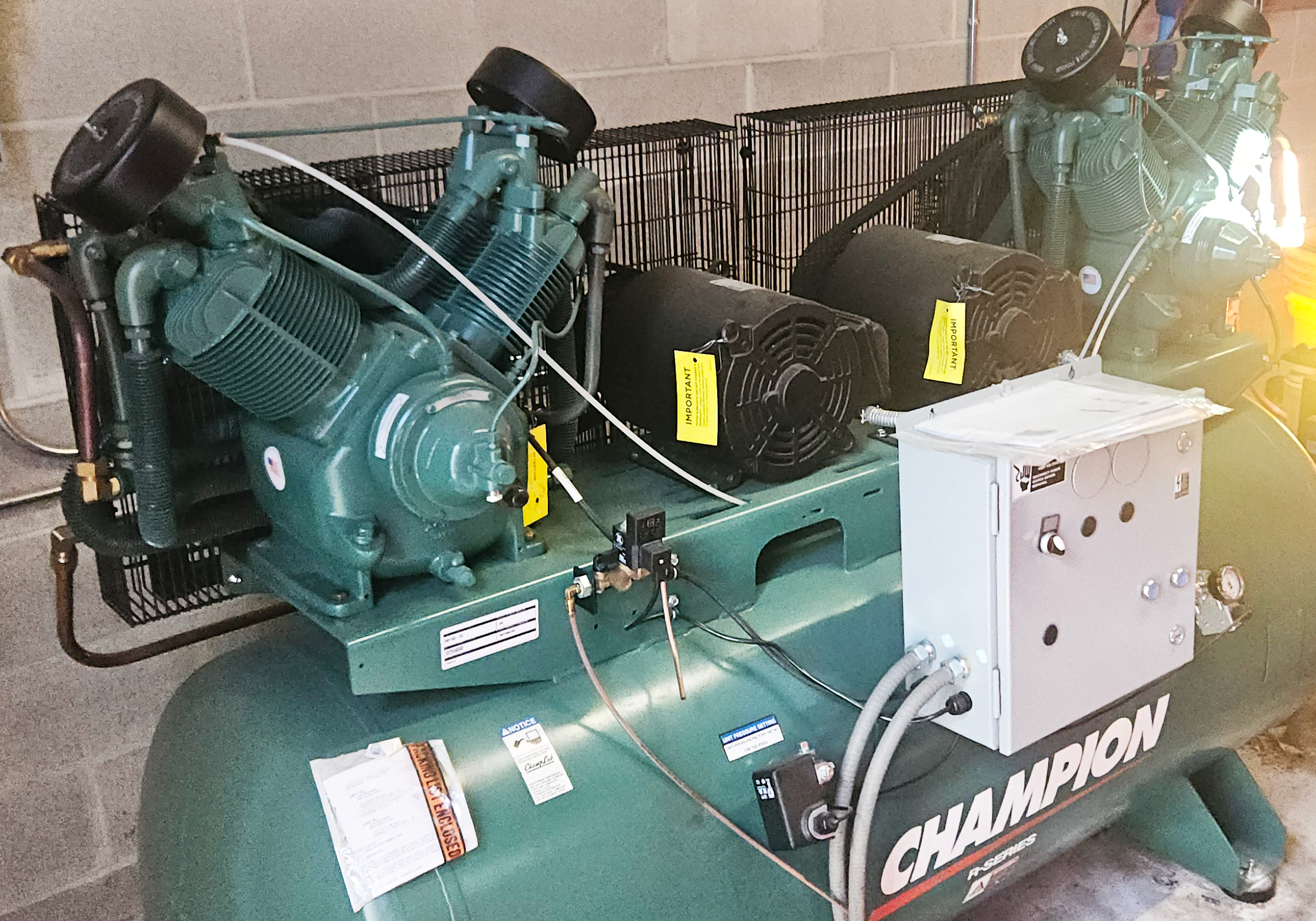 A large green Champion air compressor with dual motors and control panel sits on concrete floor by a cinder block wall in an industrial setting.