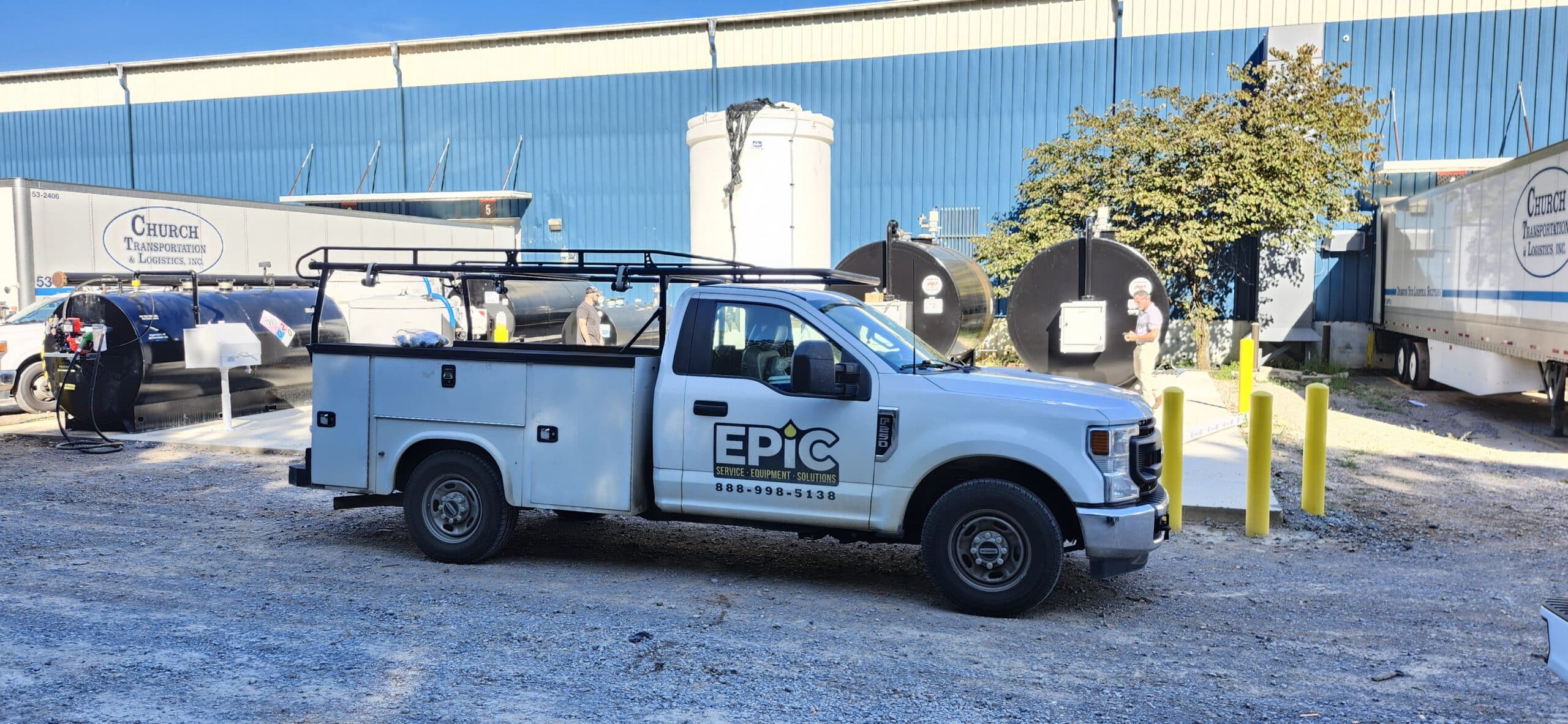 A white EPIC Service truck sits on a gravel lot by industrial tanks and trailers, with a blue warehouse and a worker in safety gear nearby.