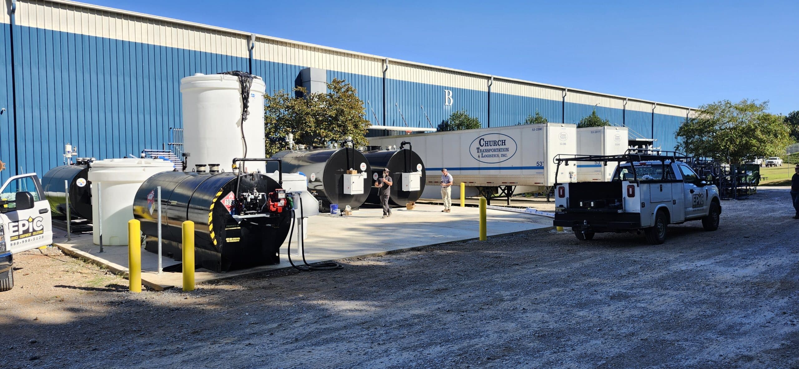 Outdoor industrial site with large fuel tanks, white van, truck, workers in safety vests by blue warehouse under clear sky; yellow bollards.