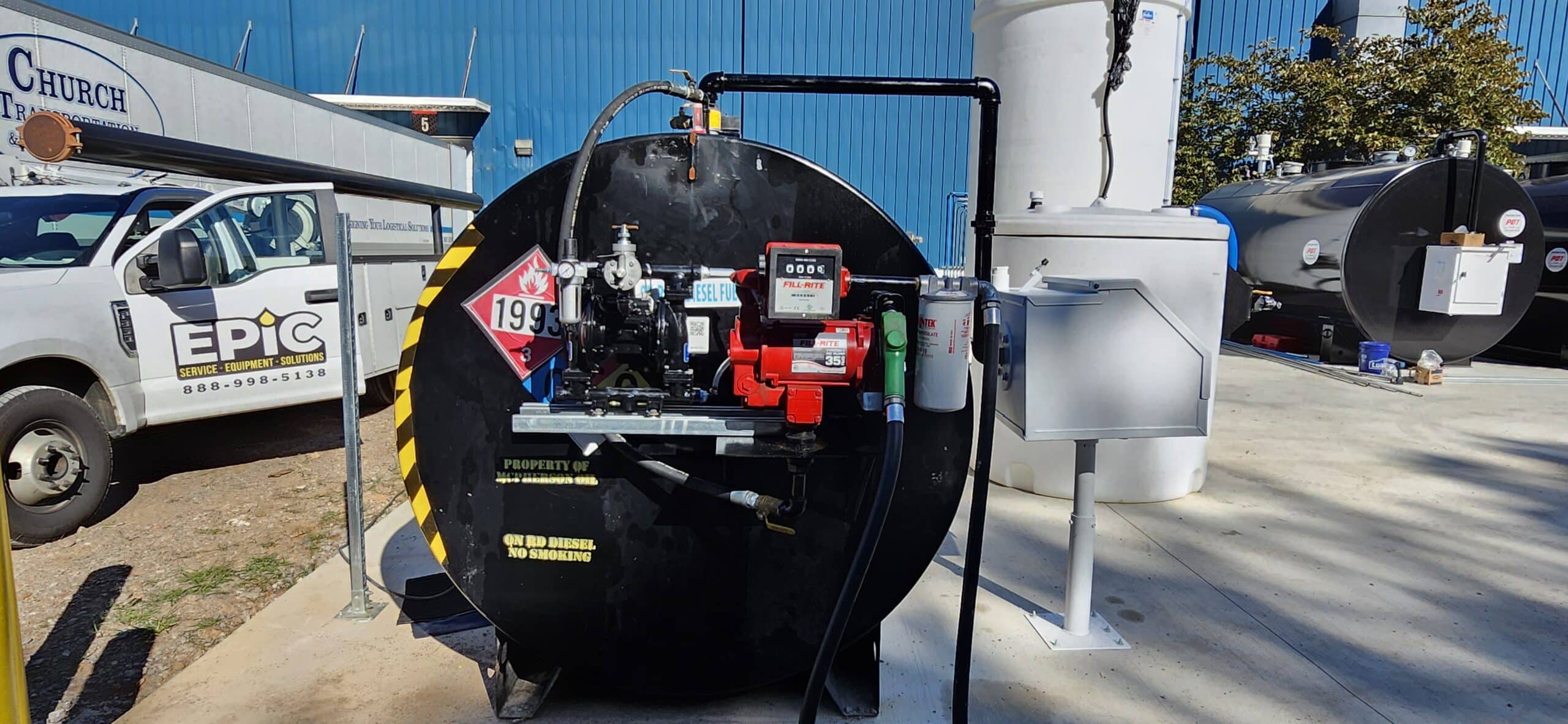 Large black fuel storage tank with hazard labels and equipment sits on concrete by a white truck and blue industrial building.
