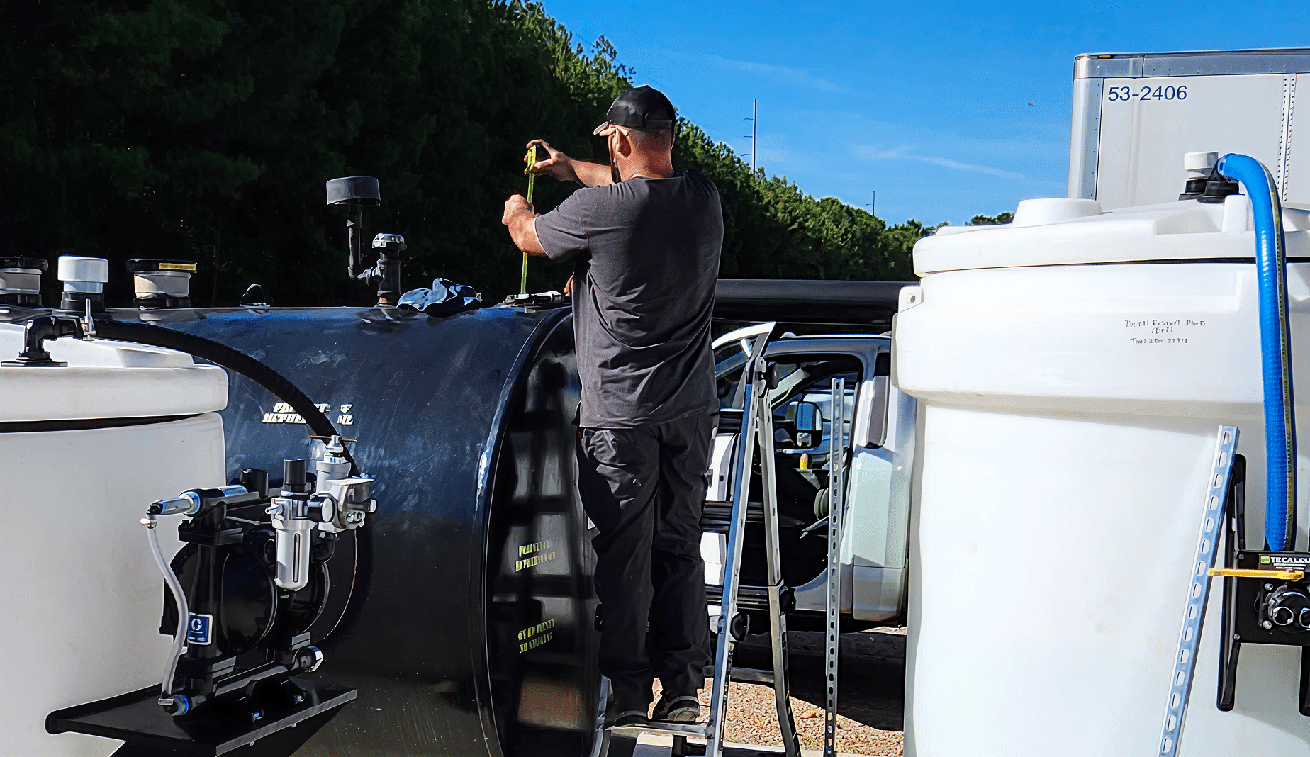 A person on a metal ladder measures a large black tank outdoors, with two white tanks and a white vehicle under a blue sky nearby.