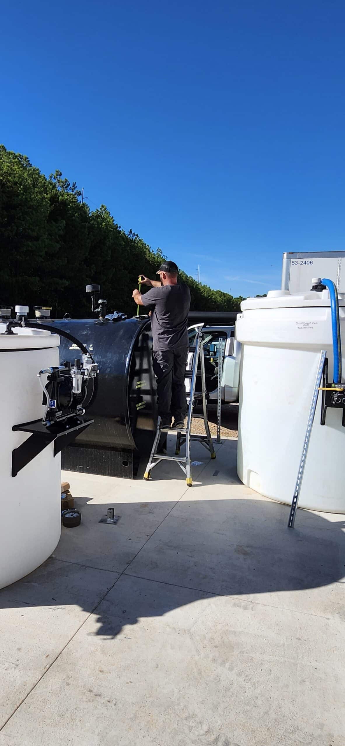 A person on a ladder works atop a large black outdoor tank, surrounded by white tanks and equipment, with trees and blue sky behind.