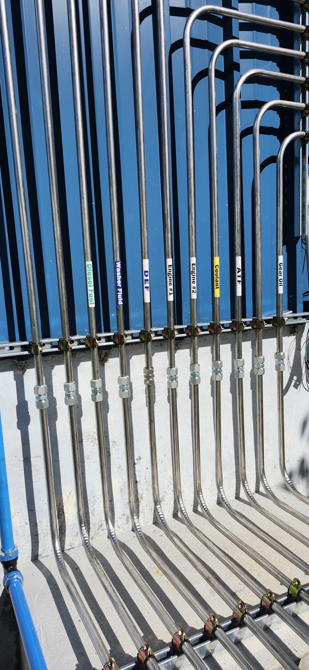 Eight shiny metal pipes run vertically along a blue wall, each labeled with colored tags like Potable Water, Flush Water, DLF, and others. Their shadows are cast on the ground below.