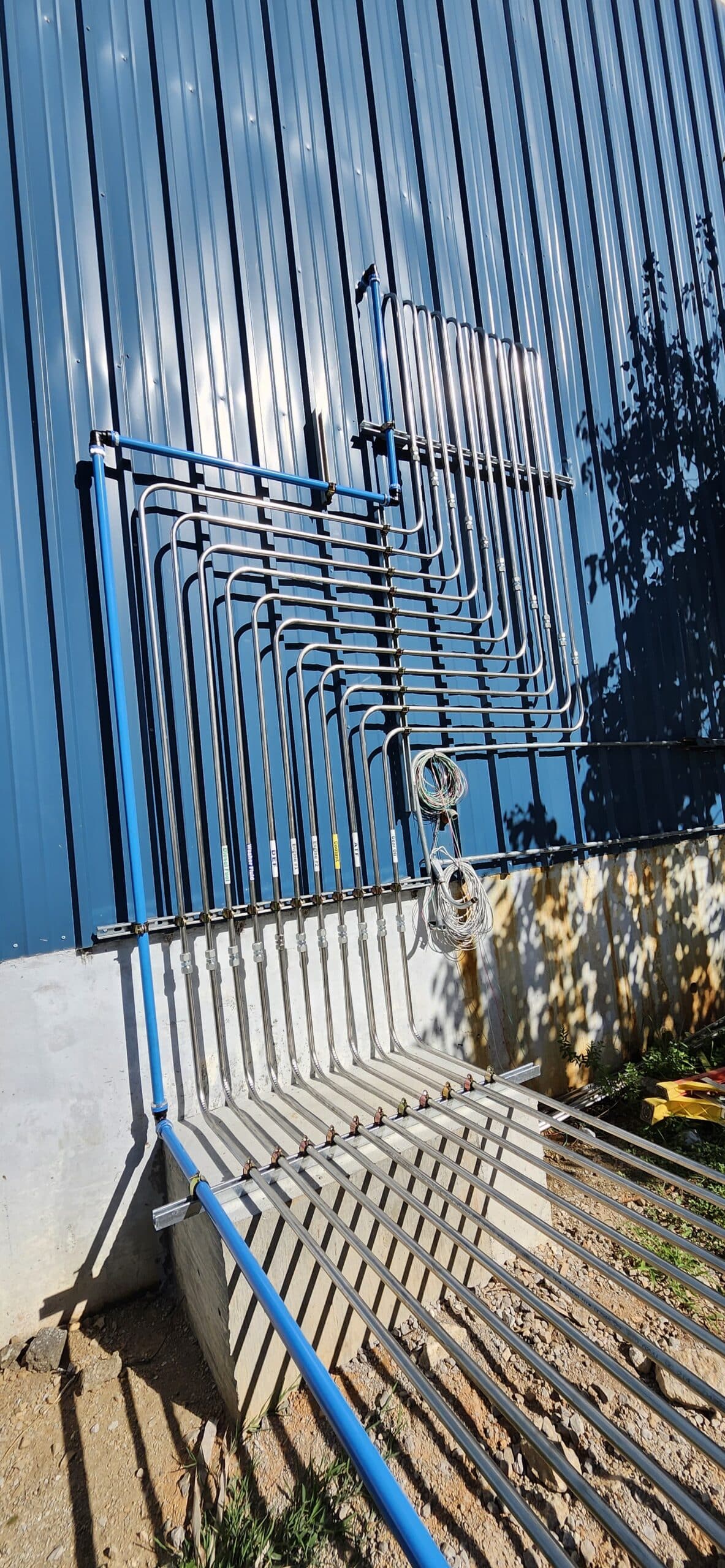 Multiple parallel black pipes with connectors run vertically and horizontally on a blue corrugated metal wall, casting intricate shadows.