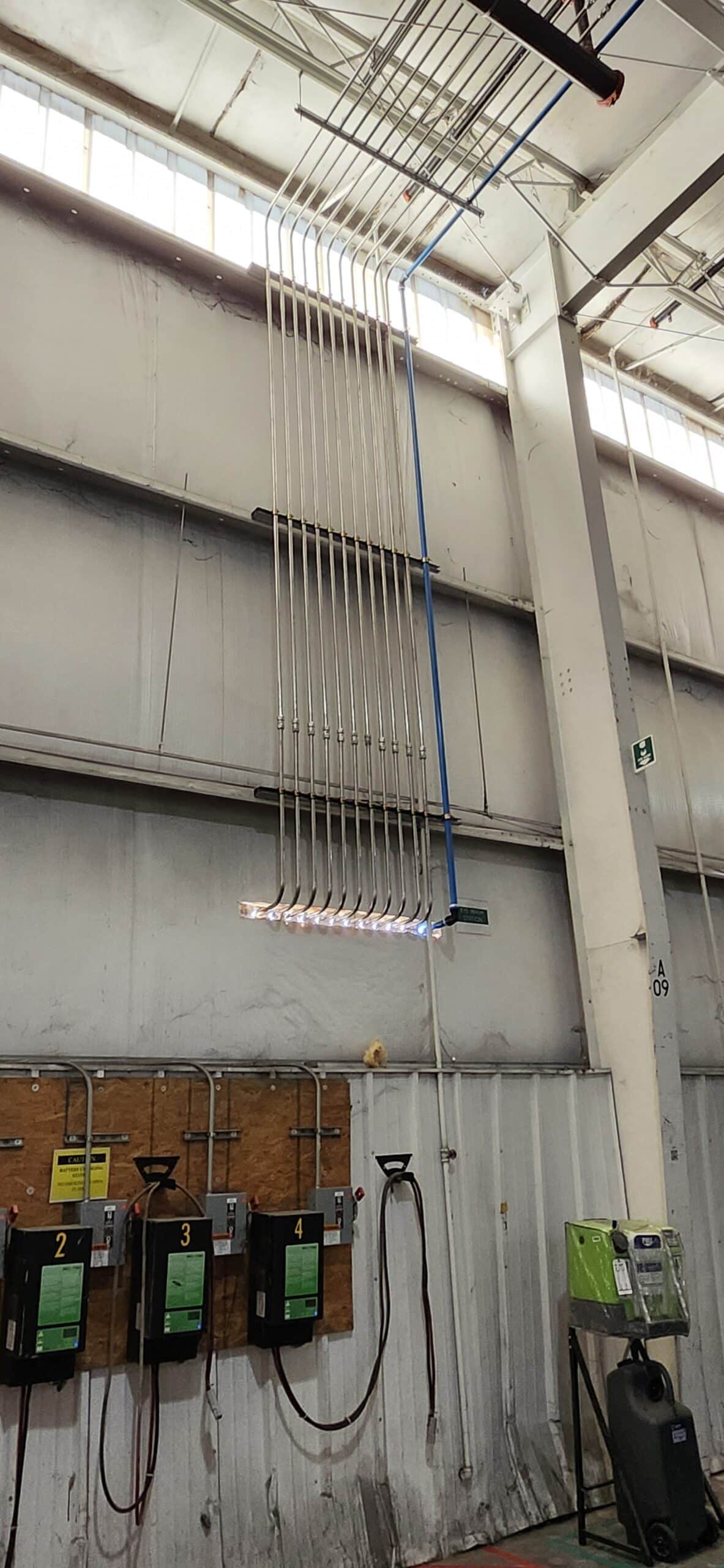 Industrial wall with ten vertical metal conduits, above four battery chargers on a wooden board, inside a large warehouse space.