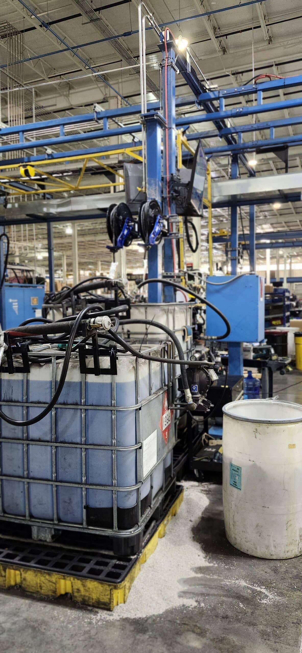 A large industrial tank with hoses rests on a pallet in a factory, surrounded by equipment, pipes, and barrels under bright lights.