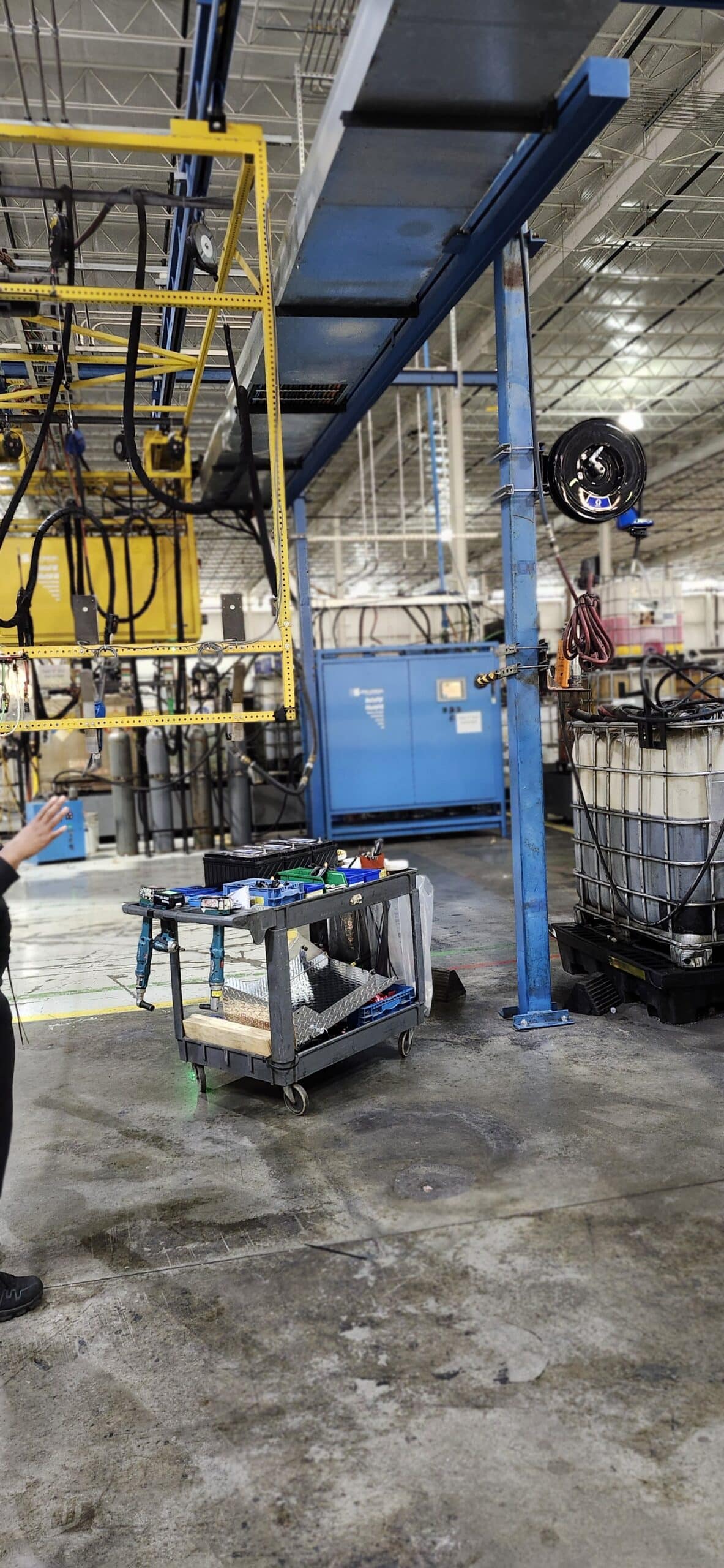 A factory floor with metal pipes, cables, a tool cart, a blue control box, and liquid container on a wet, shiny floor. Person partly visible.