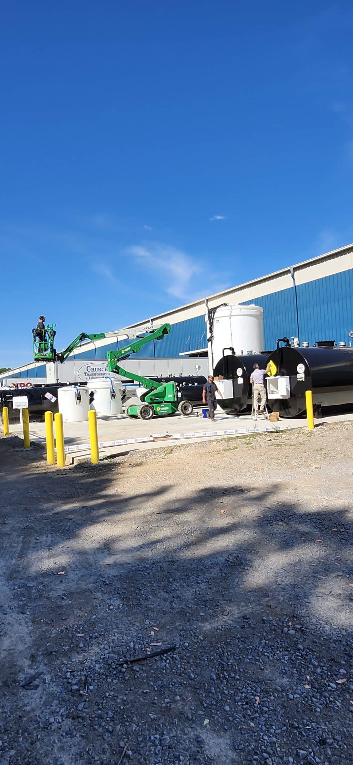 Workers operate near large black and white tanks and a green forklift outside a blue warehouse, with yellow bollards lining the front.