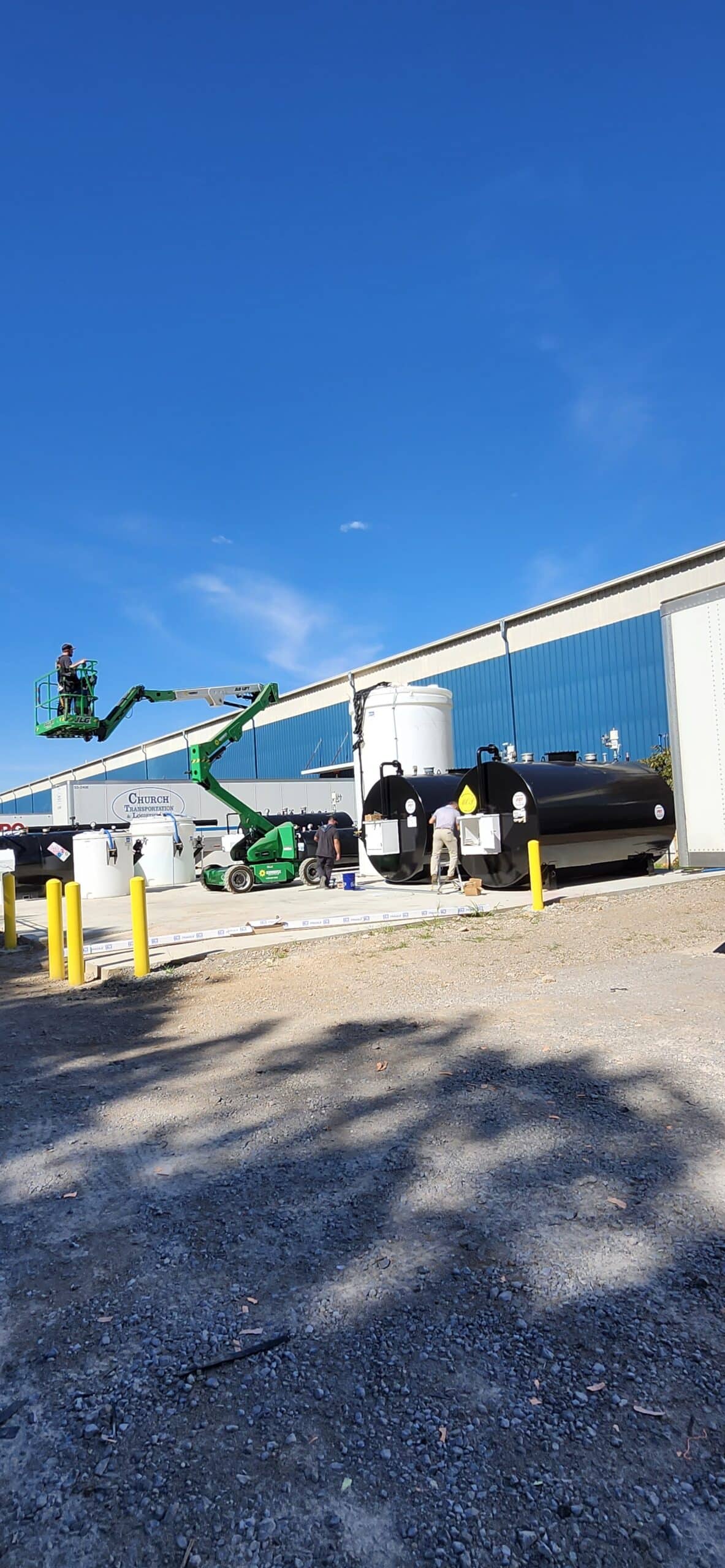 Workers in hard hats operate cherry pickers by large industrial tanks outside a building with blue windows; yellow bollards in front.