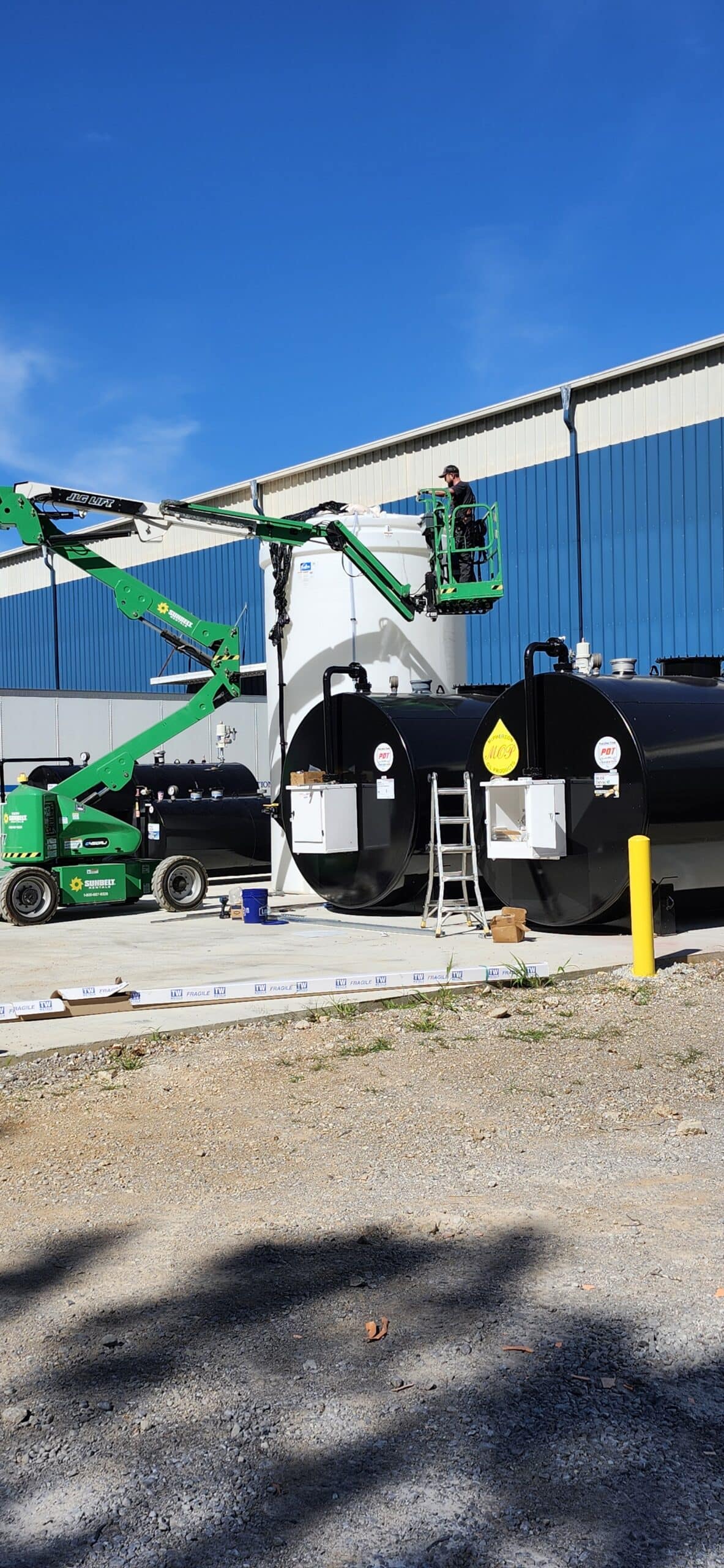 Two workers in green lifts work atop large black cylindrical tanks outside a blue-and-white industrial building under a clear sky.