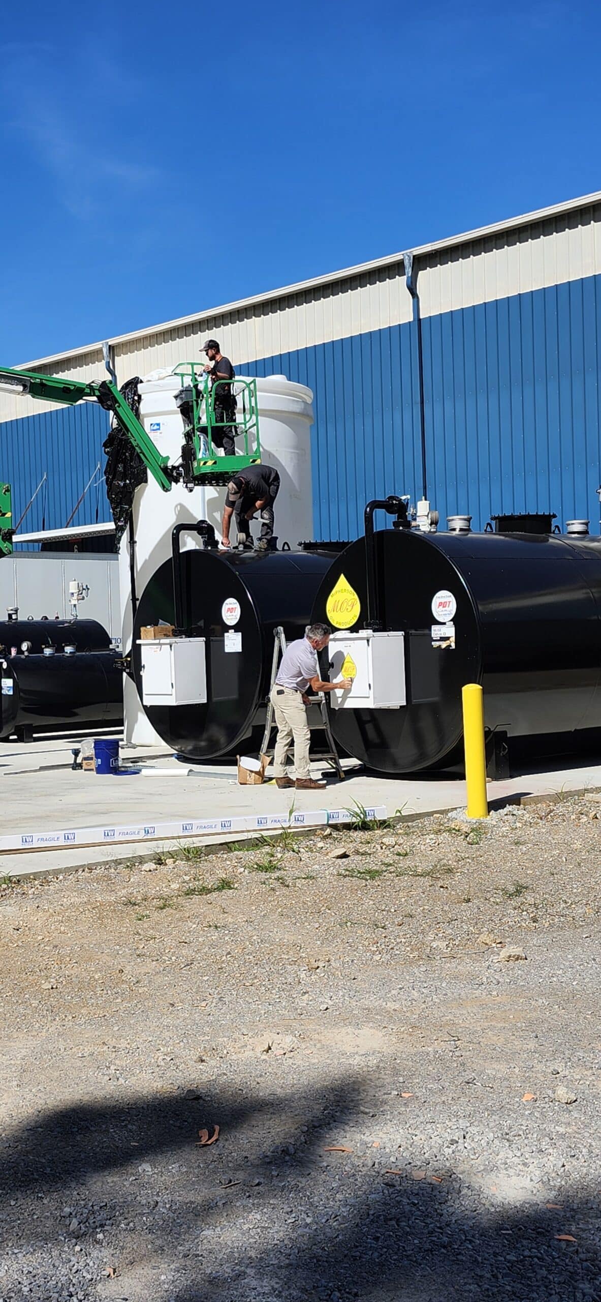 Two workers on a lift inspect black storage tanks outside an industrial building as another applies a yellow label under a blue sky.