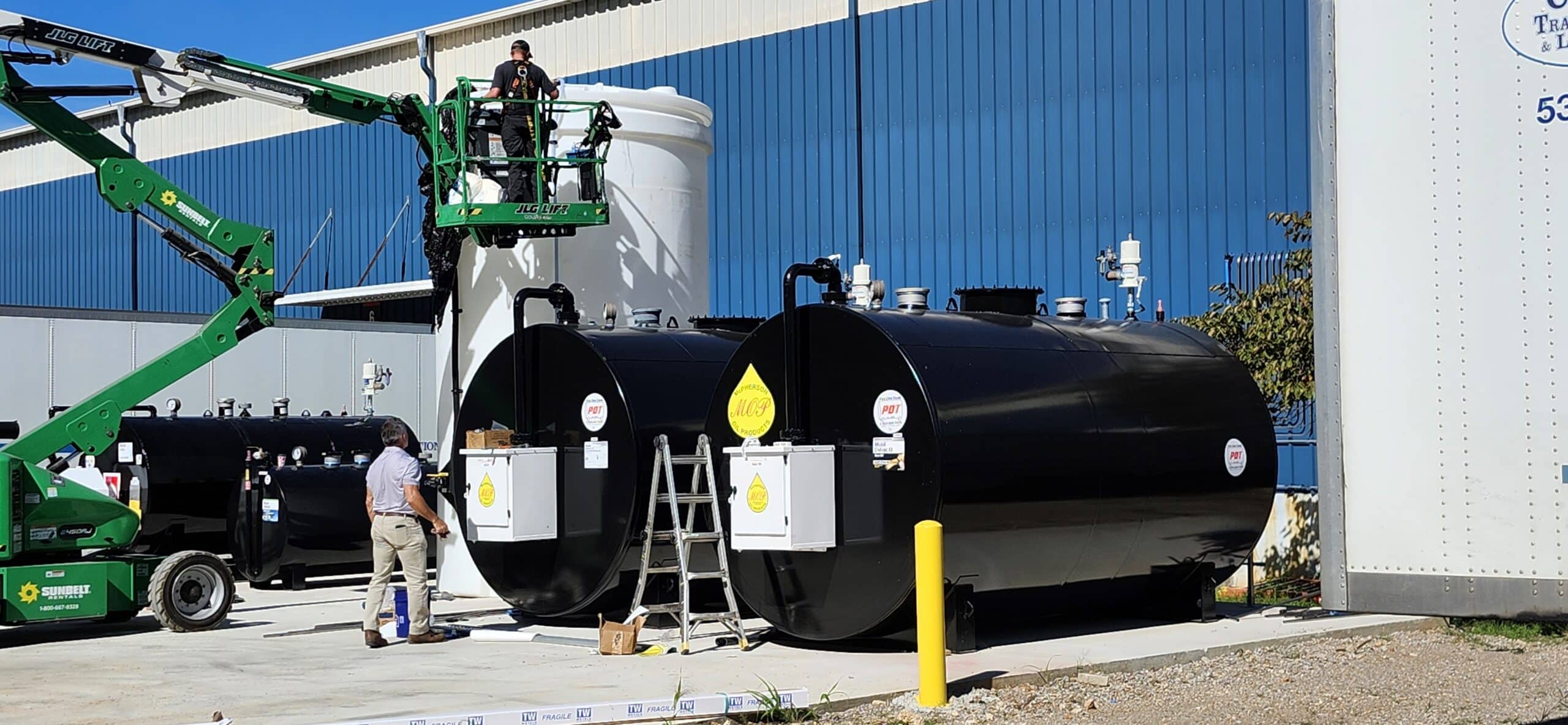 Two workers use a green boom lift and a ladder to reach the tops of large black fuel tanks outside an industrial building. Equipment nearby.