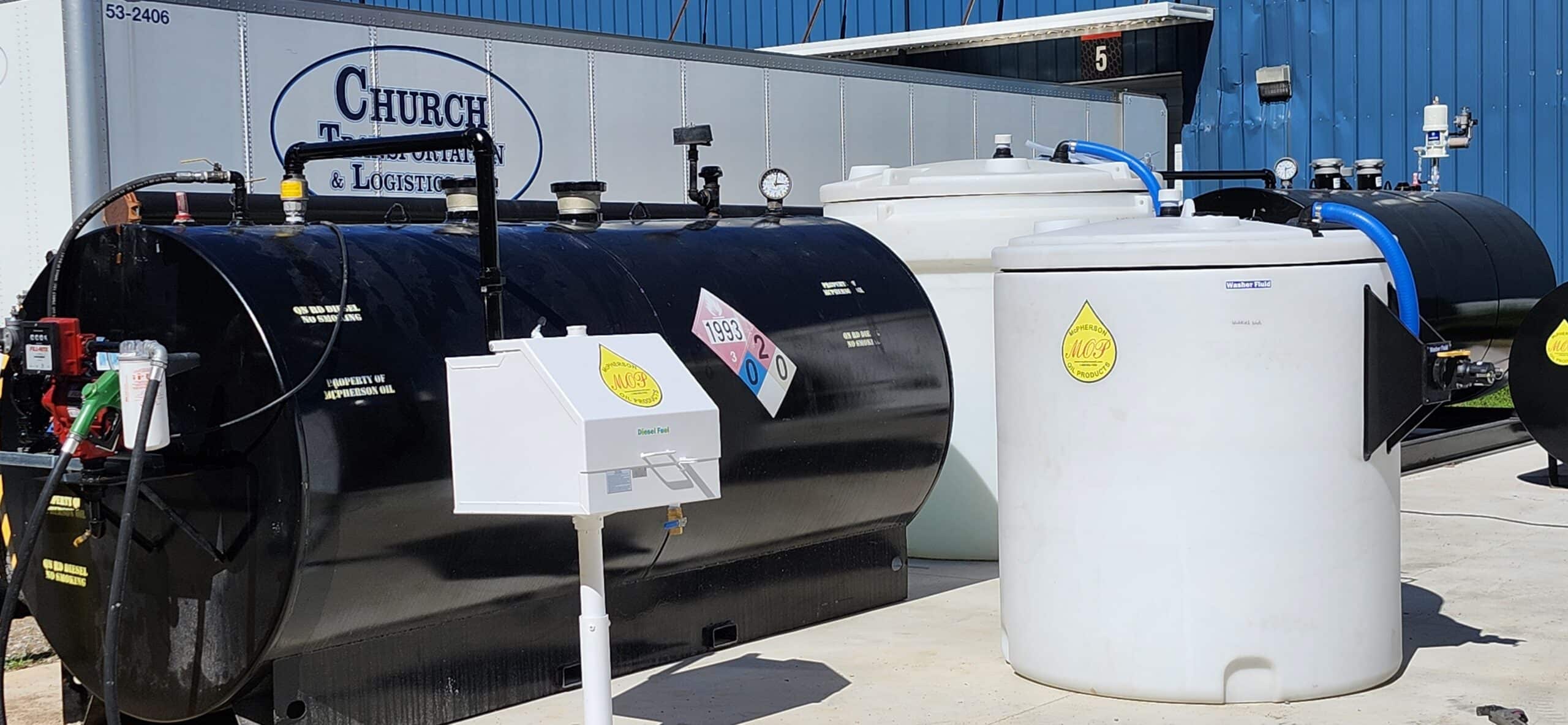 Several black and white storage tanks stand outdoors by a blue industrial building and a Church logistics trailer, with hoses and warnings.
