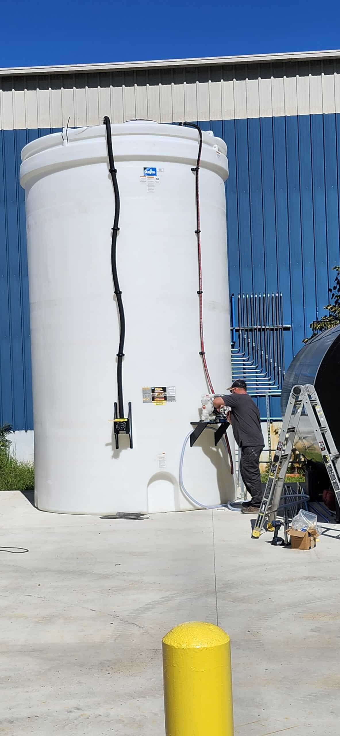 A person by a large white tank with black and red pipes, using a hose; ladder, tools, and blue building under a clear sky nearby.